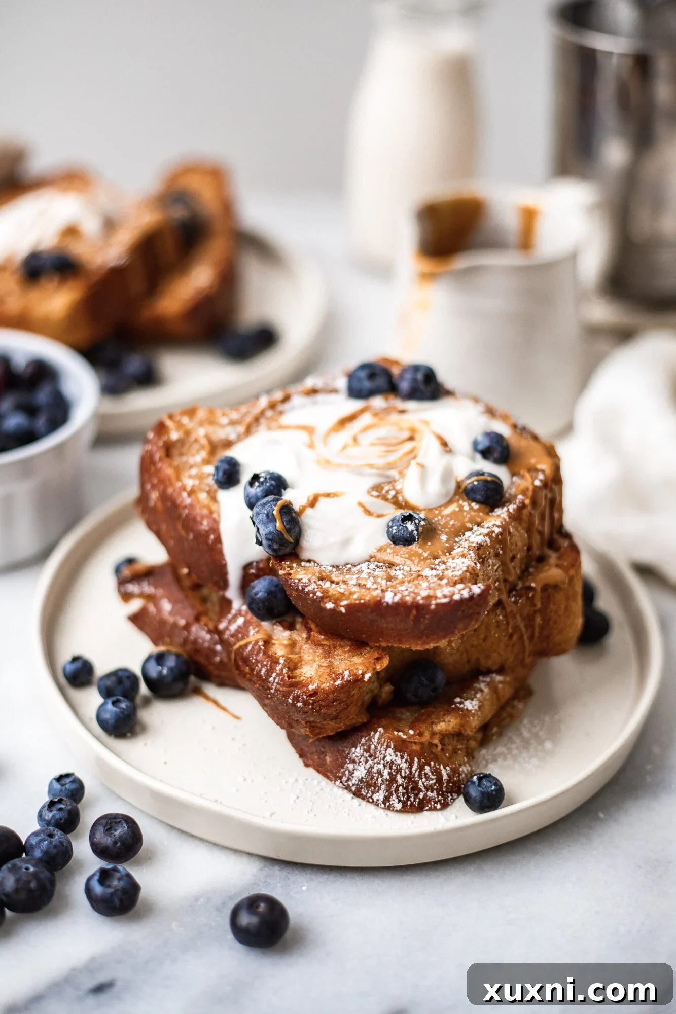 stack of vegan French toast with blueberries on top next to a jar dripping with peanut butter and a glass of coconut milk.