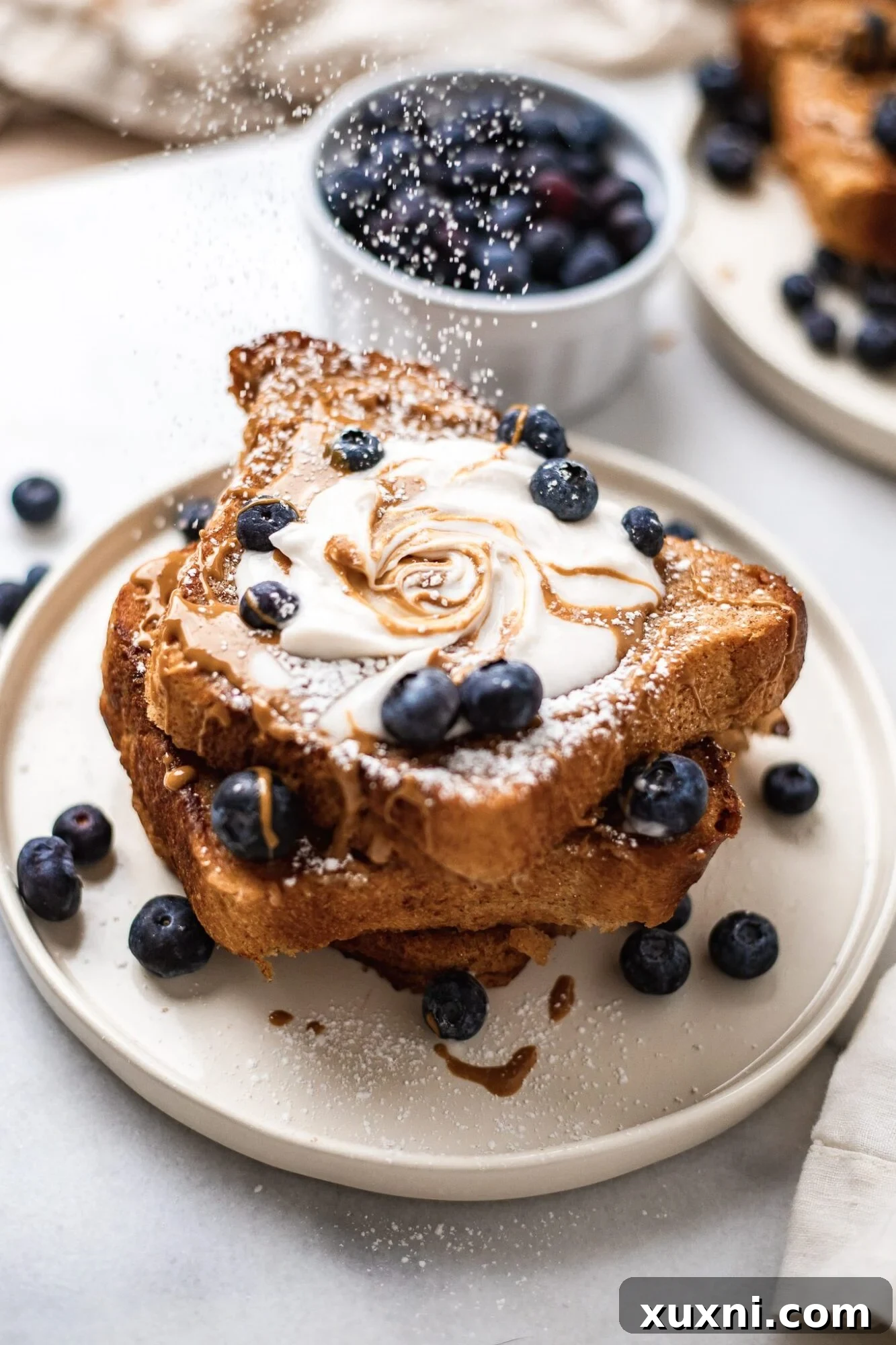 powdered sugar being sprinkled onto vegan French toast that is topped with blueberries, coconut whipped cream, maple syrup, and peanut butter. Bowl of blueberries is next to it.