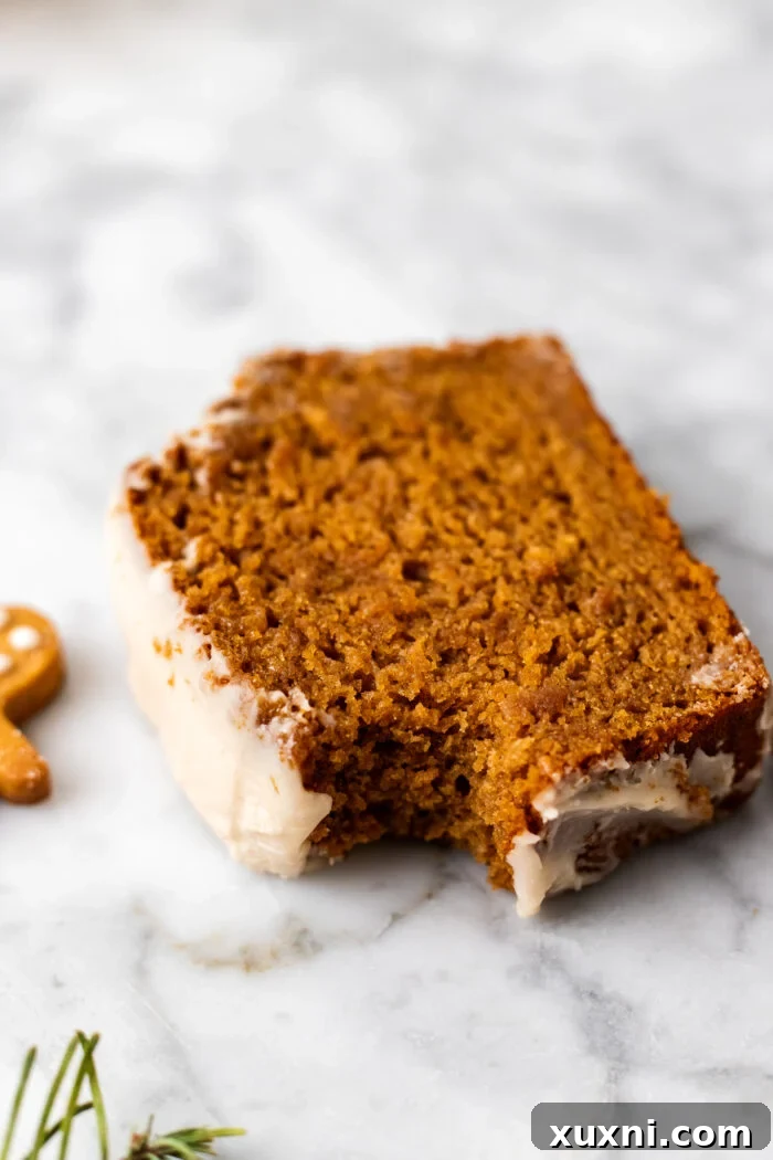 A close-up of a bitten slice of vegan gingerbread loaf cake, showing the moist interior and the creamy cashew maple frosting, resting on a marble surface.