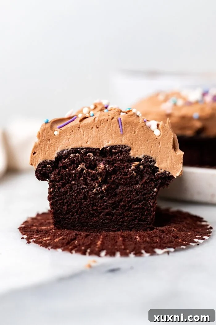 Two moist and fluffy vegan gluten-free chocolate cupcakes, one sliced in half to reveal its tender crumb, topped with dairy-free chocolate sour cream buttercream and sprinkles.