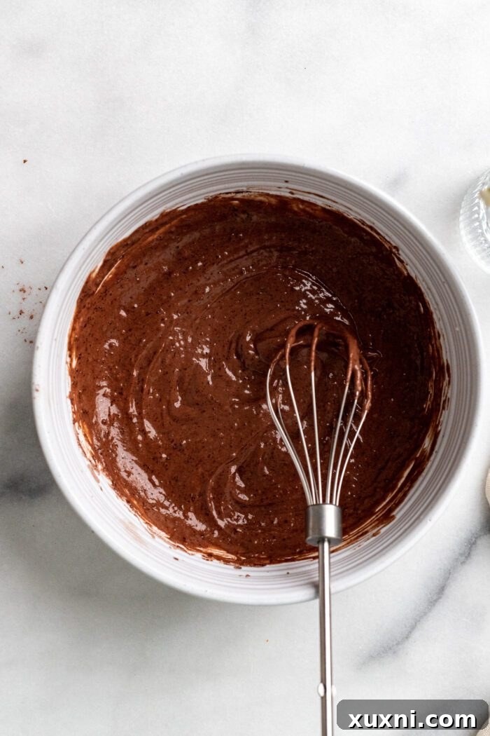 Wet ingredients being mixed into dry ingredients to form a smooth chocolate cupcake batter in a glass bowl.