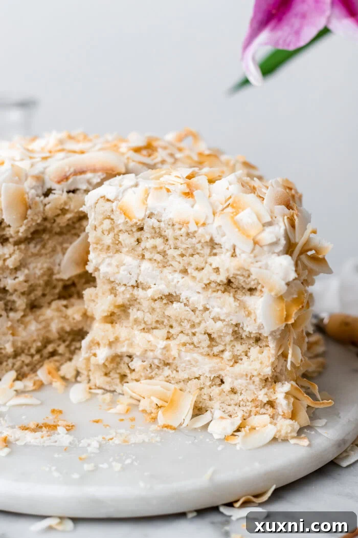 A slice of vegan coconut cake, generously covered in dairy-free coconut cream cheese frosting and toasted coconut, on a cake plate.