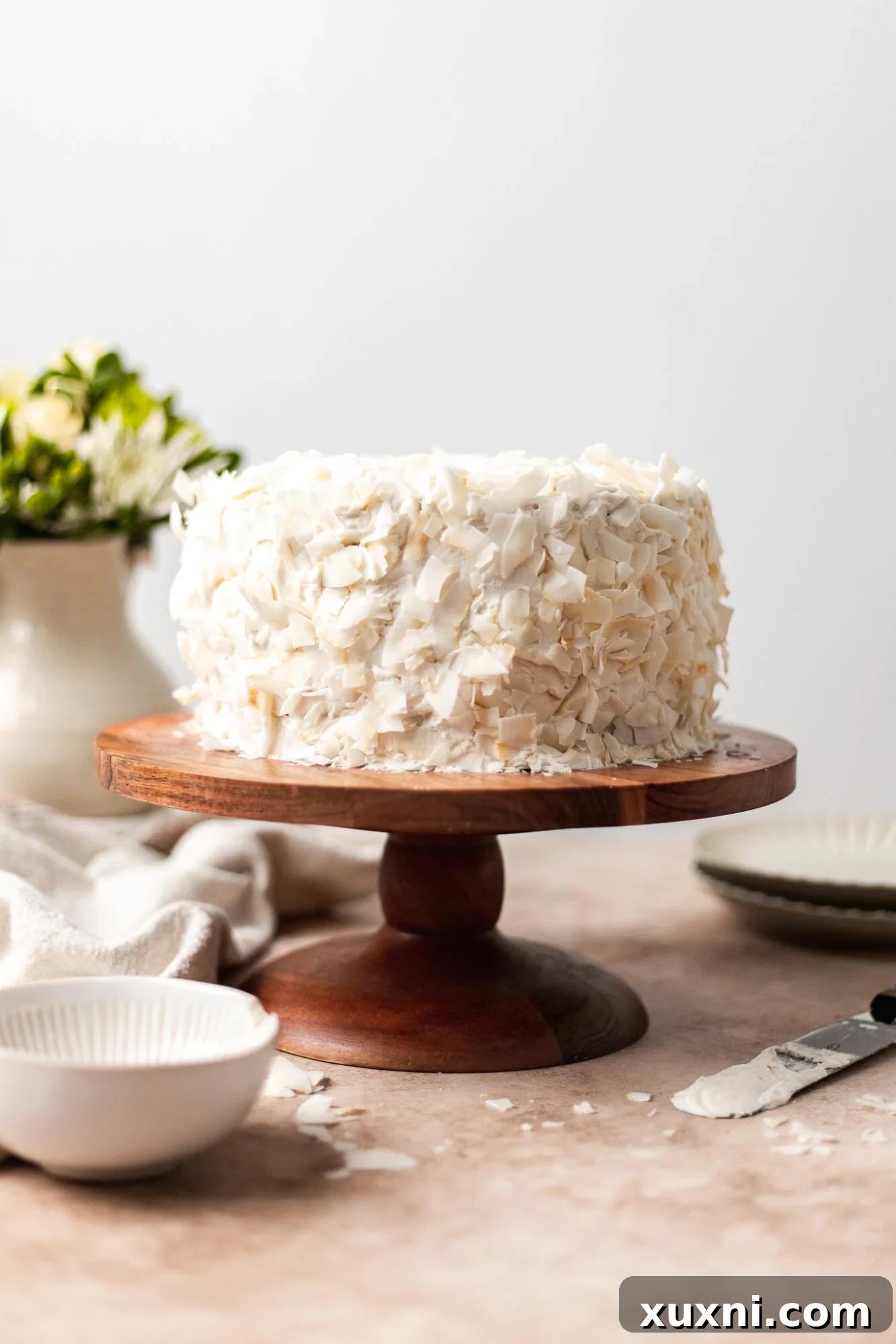A fully frosted and decorated vegan coconut cake, topped with a generous amount of toasted coconut flakes.