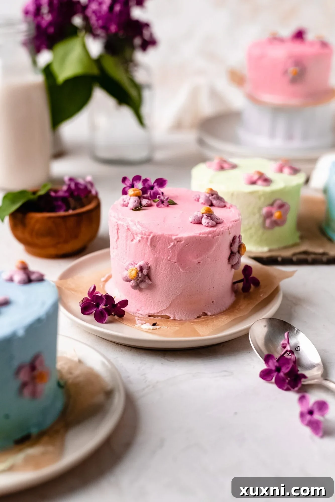 Three mini cakes, each with different decorations: a vanilla cake with flowers, a pistachio cake with nuts, and a red velvet cake with piped frosting.