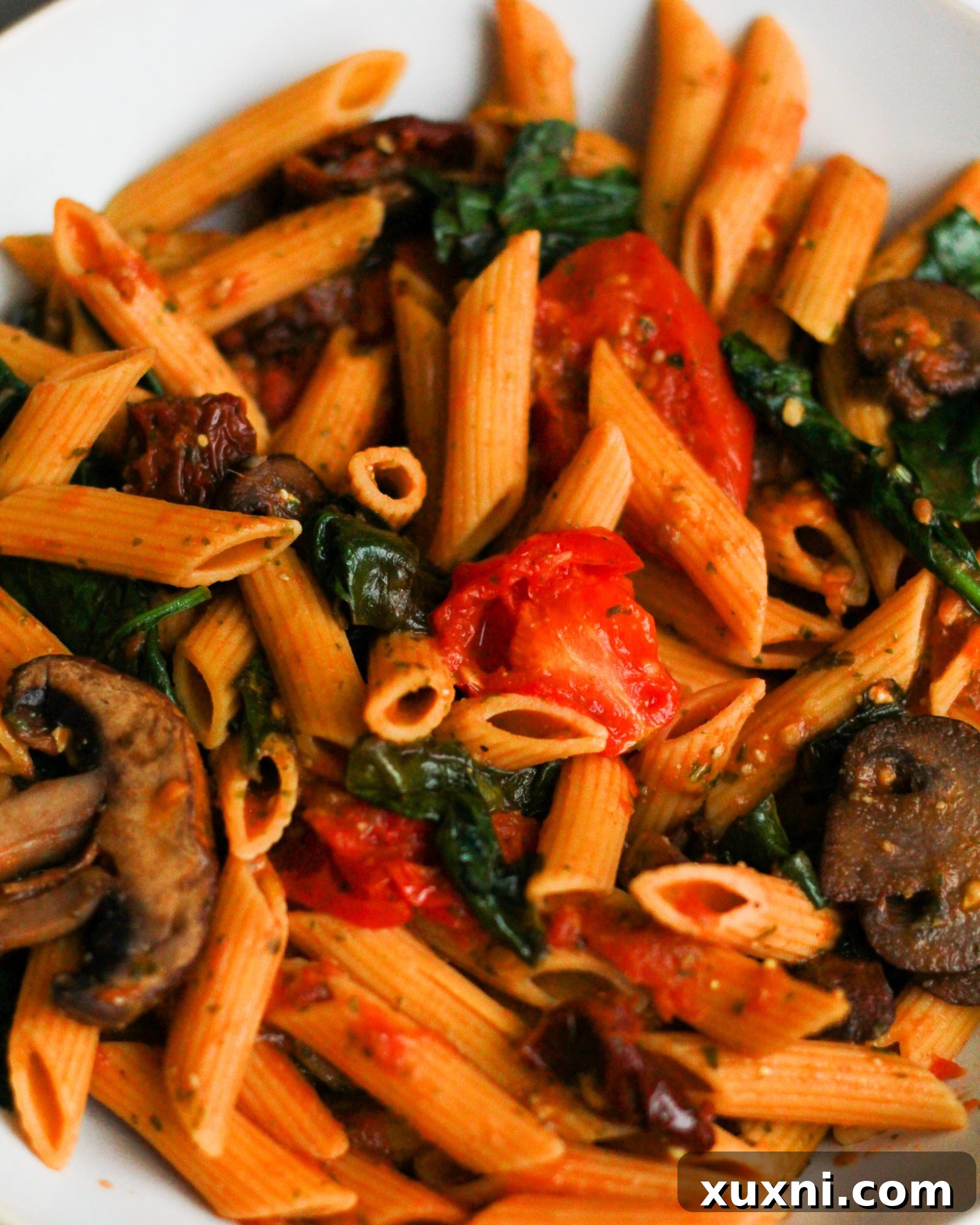 Assortment of fresh ingredients for vegan pesto pasta, including colorful tomatoes, spinach, mushrooms, and a jar of sun-dried tomatoes.