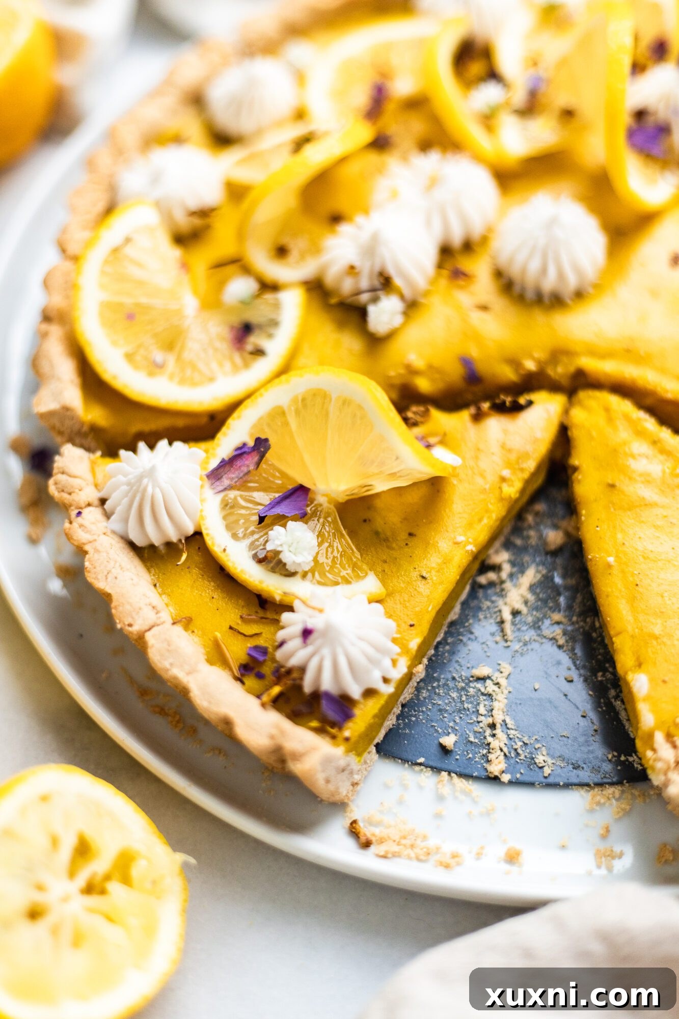 A single slice of vegan lemon tart, garnished with lemon zest and edible flowers, on a white plate.