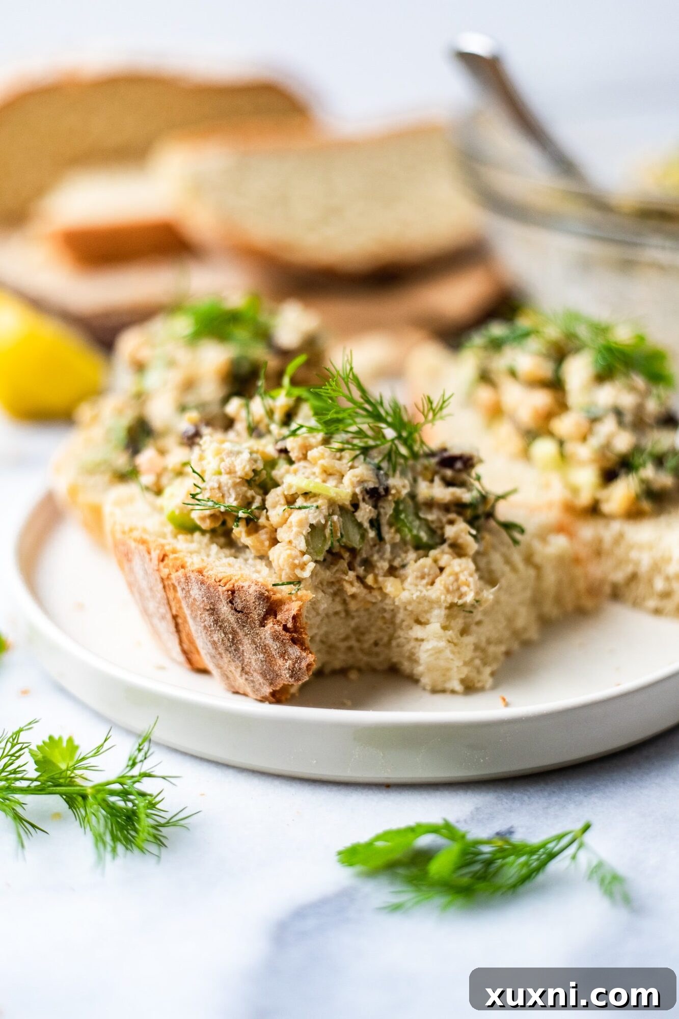 Plant Based Chickpea Tuna Salad 2 Delicious vegan chickpea tuna salad served on a slice of bread, garnished with fresh dill and parsley, resting on a white plate.