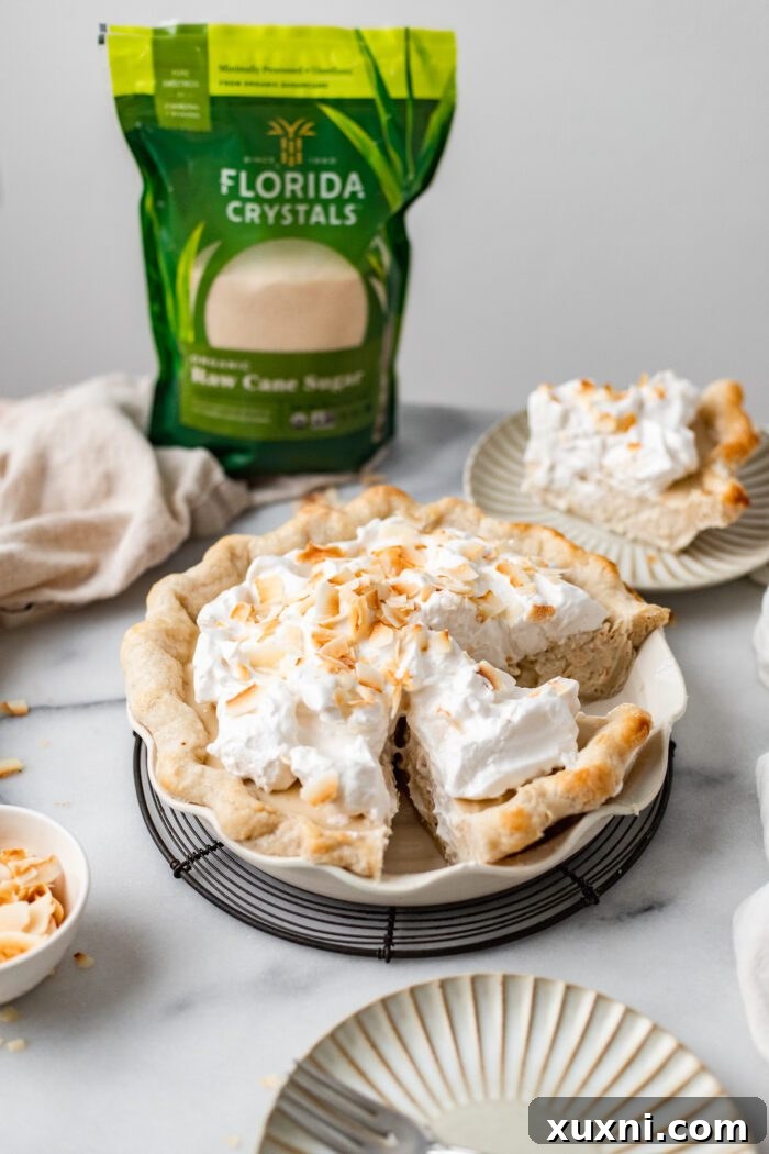 A beautifully sliced vegan coconut cream pie on a white plate, revealing its layers of flaky crust, rich coconut filling, and fluffy whipped cream, with more slices in the background.
