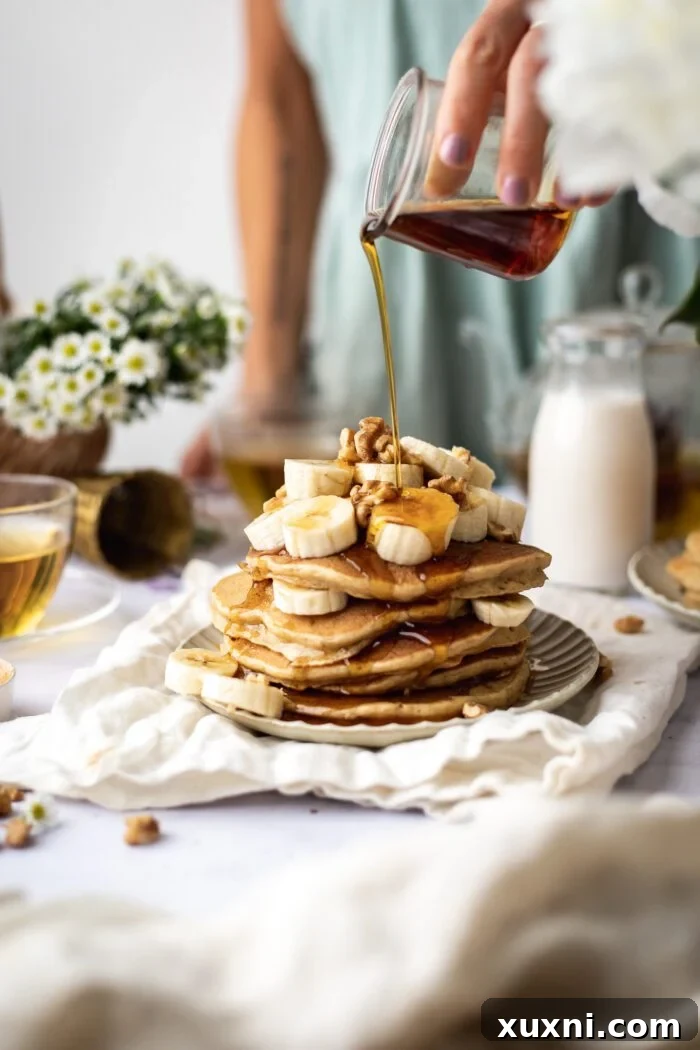 stack of delicious vegan banana pancakes with a hand pouring maple syrup over them, highlighting their fluffy texture