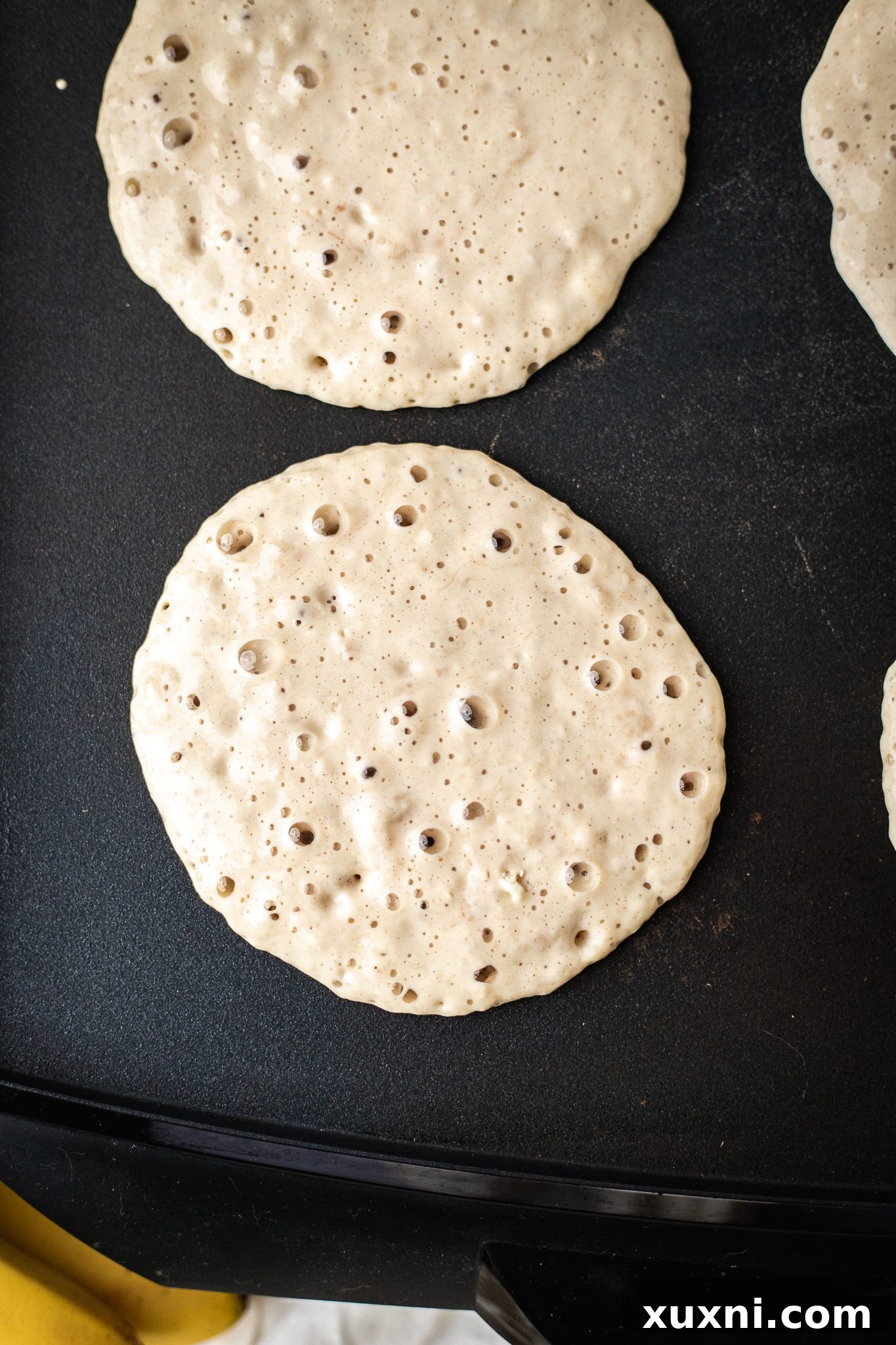 vegan banana pancakes being cooked on a pancake griddle, with bubbles forming on the surface