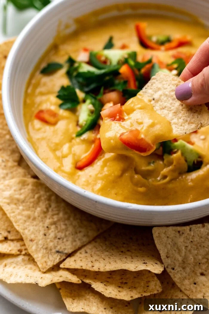 A hand dipping a tortilla chip into a bowl of vegan queso, showcasing its creamy texture.