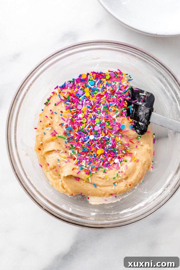 Colorful vegan sprinkles being added to the prepared cookie dough in a mixing bowl.