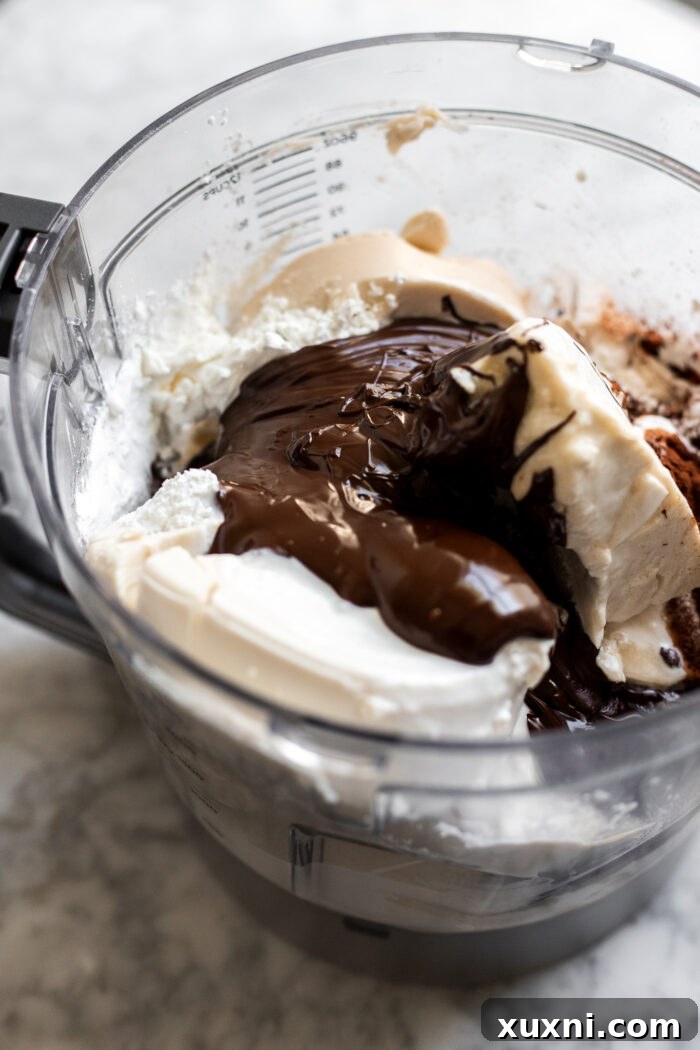 Vegan cheesecake filling ingredients, including vegan cream cheese, yogurt, and melted chocolate, being blended in a food processor.