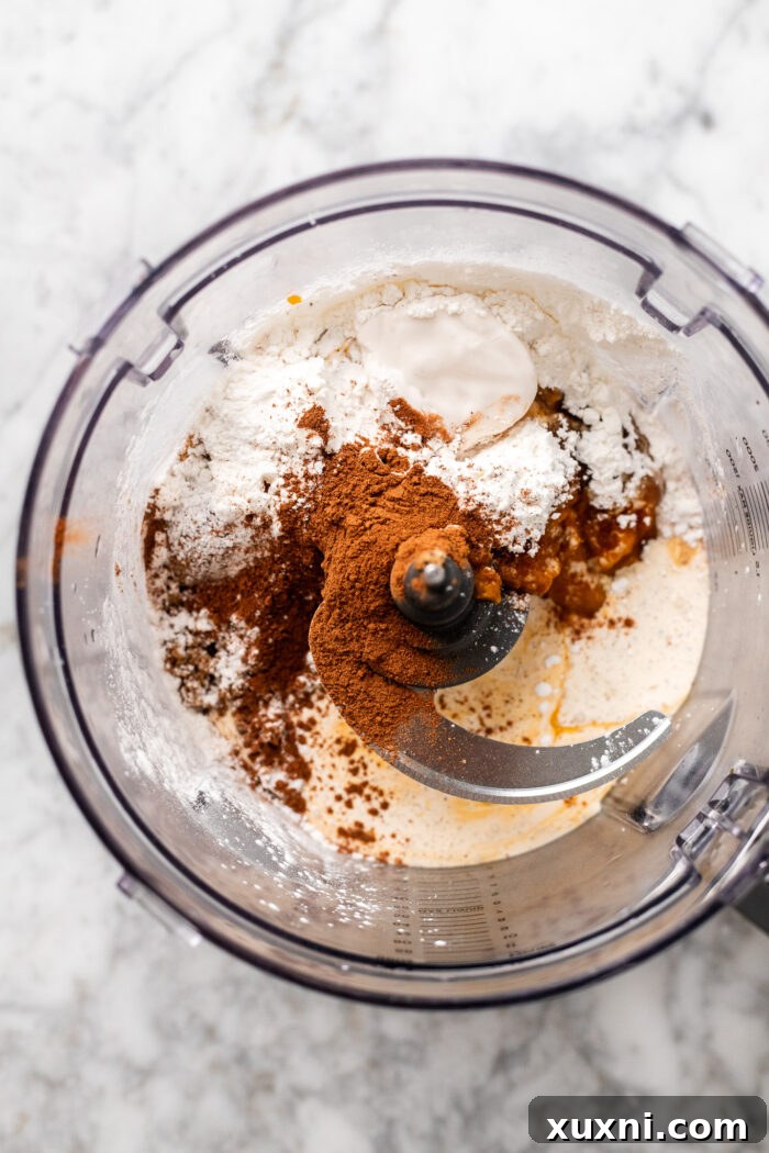All pumpkin pie filling ingredients, including pumpkin puree and vegan eggnog, gathered in a food processor, ready for blending.