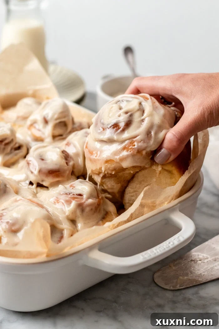 A hand reaching into a baking dish to grab a perfectly iced vegan cinnamon roll.