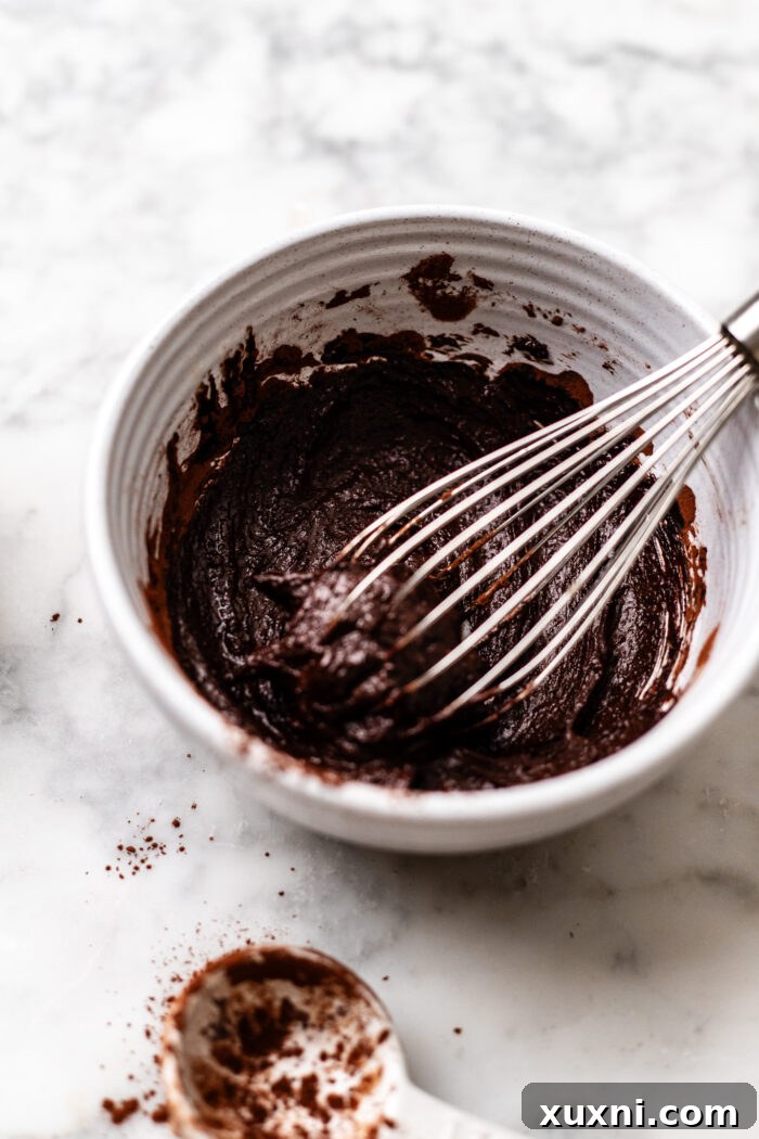 Ingredients for fudge ripple sauce, including cocoa powder and vegan cream, being whisked together in a bowl until smooth.