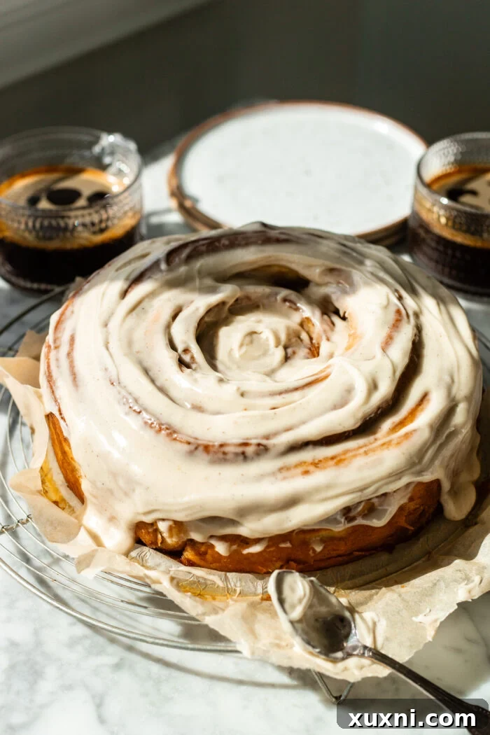 Frosted giant vegan cinnamon roll cake with cups of coffee on a marble table, ready to be sliced and enjoyed.