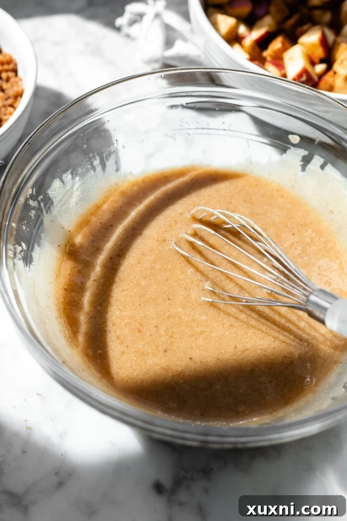 Hands whisking wet ingredients including browned vegan butter, sugars, and dairy-free yogurt in a large bowl to create the cake batter base.