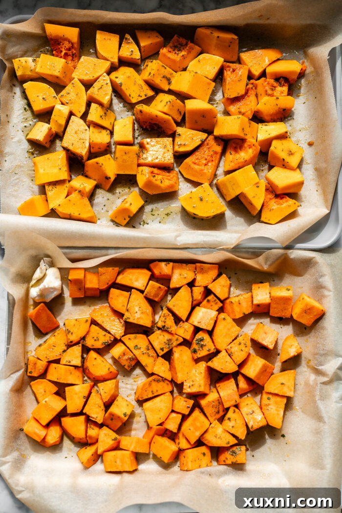Cubed butternut squash, sweet potato, sliced onion, and a garlic bulb seasoned with olive oil and spices on a parchment-lined baking sheet.
