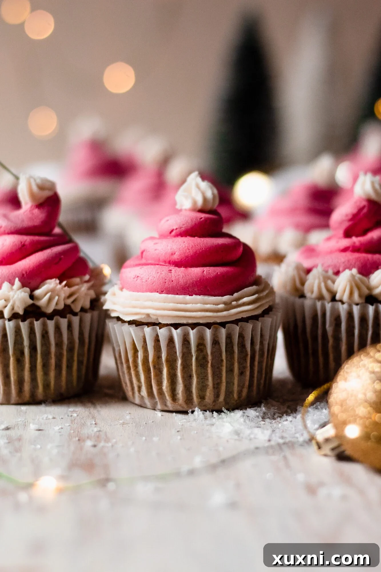 Festive Vegan Pistachio Santa Hat Cupcakes 8 A charming line-up of green pistachio Christmas cupcakes, each topped with a red and white Santa hat frosting, illuminated by soft string lights.